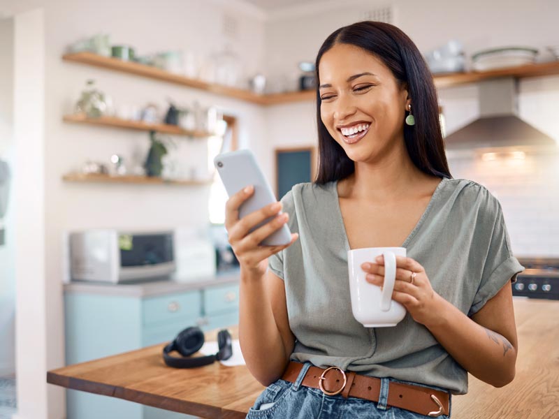 Happy young woman standing in her kitchen at home and using smartphone while drinking a cup of coffee